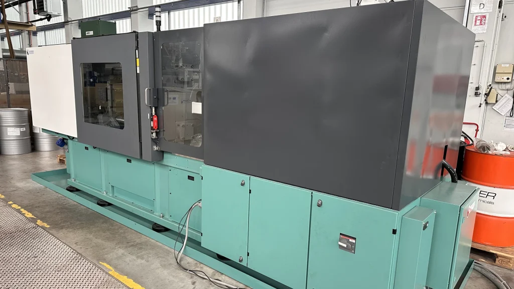 Eine große, grün-graue Industriemaschine steht in einer Fabrikumgebung, mit Kabeln am Boden und einem Sichtfenster auf der linken Seite. Im Hintergrund sind verschiedene Geräte und Fässer zu sehen.