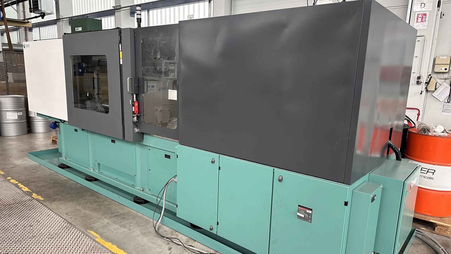 Eine große, grün-graue Industriemaschine steht in einer Fabrikumgebung, mit Kabeln am Boden und einem Sichtfenster auf der linken Seite. Im Hintergrund sind verschiedene Geräte und Fässer zu sehen.