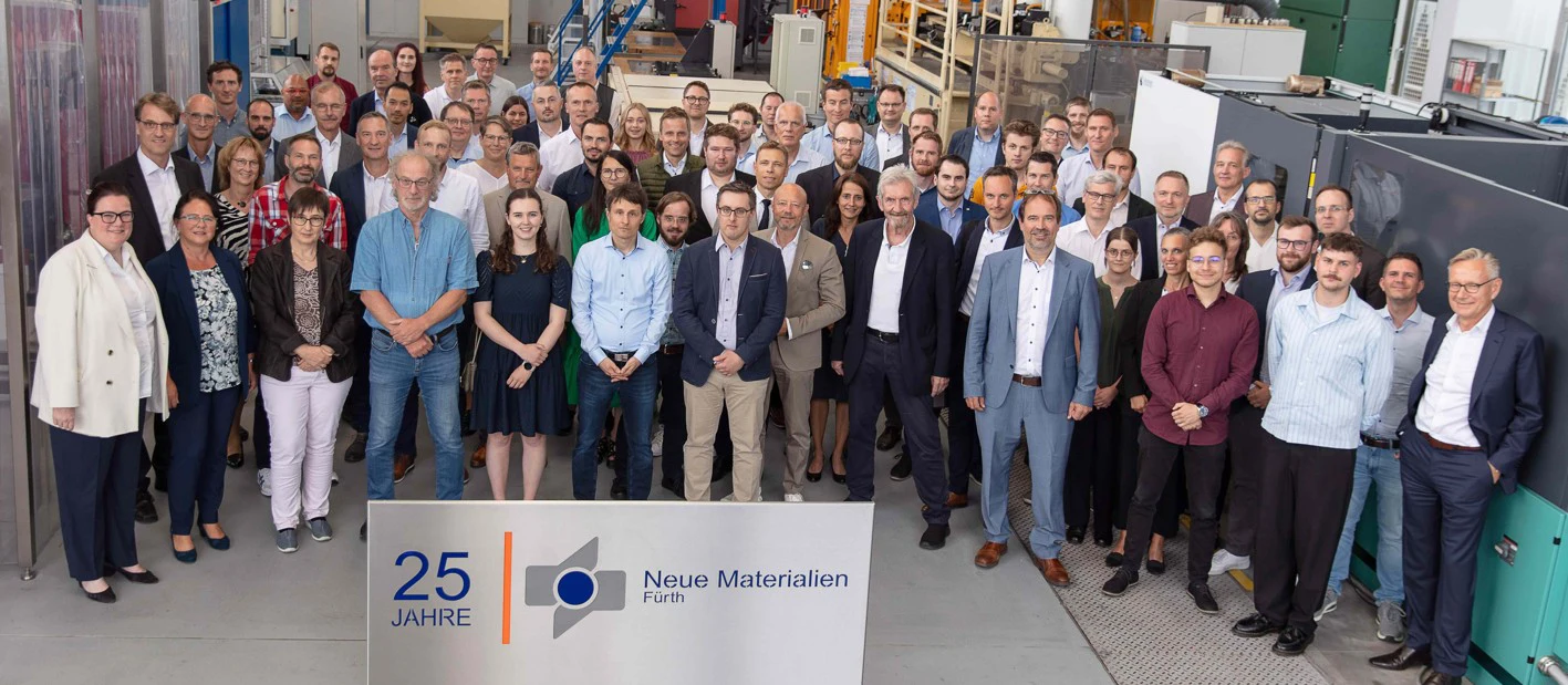 Eine große Gruppe von Männern und Frauen steht zusammen in einem industriellen Arbeitsbereich und posiert für ein Foto hinter einem Schild mit der Aufschrift "25 Jahre Neue Materialien Fürth".