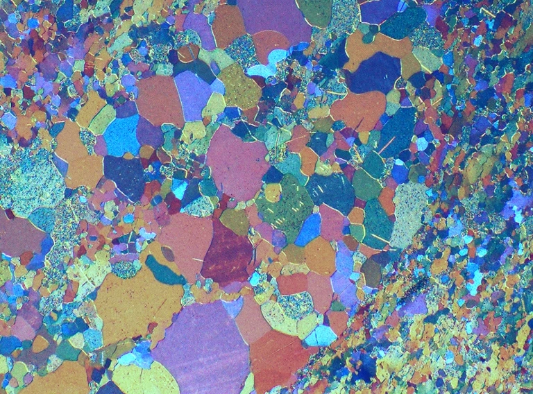 Buntes, mosaikartiges Muster aus unregelmäßigen, ineinandergreifenden Formen in den Farben Blau, Orange, Rosa, Gelb und Grün, das einem dünnen Gesteins- oder Mineralienschnitt ähnelt, der unter einem Mikroskop mit polarisiertem Licht betrachtet wird.