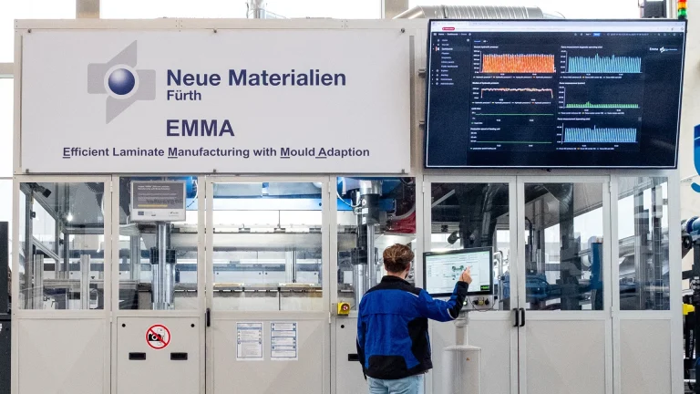 Eine Person bedient ein Bedienfeld vor einer großen Industriemaschine mit der Aufschrift Neue Materialien Fürth EMMA. Ein Bildschirm auf der rechten Seite zeigt Datengrafiken an, und der Bereich ist sauber und gut beleuchtet.