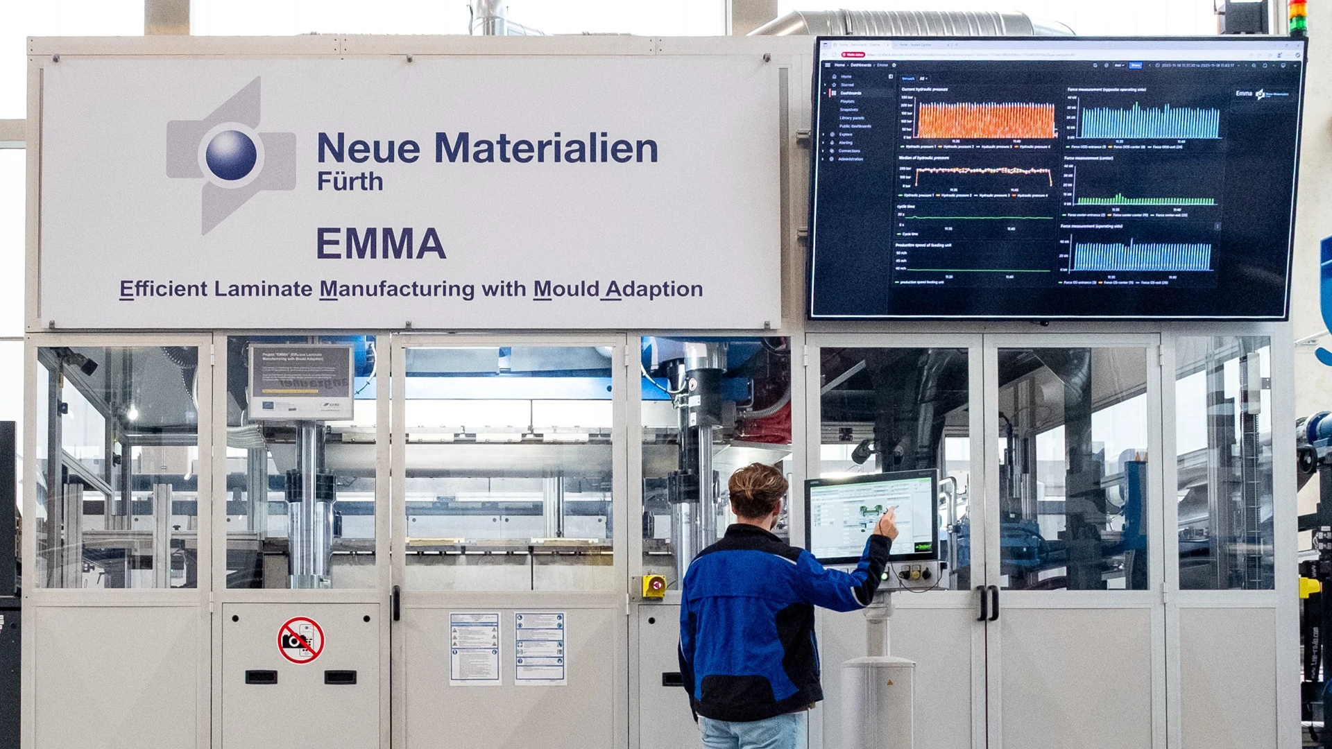Eine Person bedient ein Bedienfeld vor einer großen Industriemaschine mit der Aufschrift Neue Materialien Fürth EMMA. Ein Bildschirm auf der rechten Seite zeigt Datengrafiken an, und der Bereich ist sauber und gut beleuchtet.