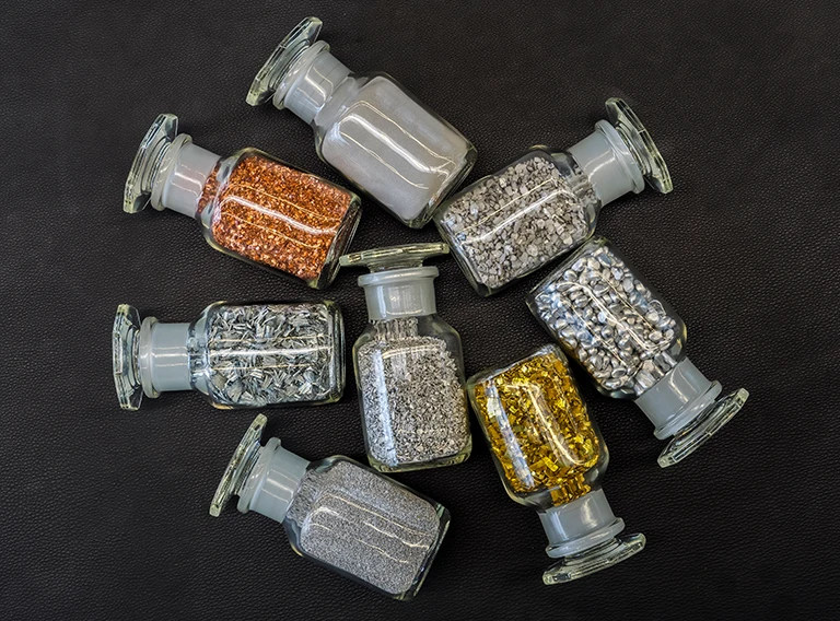 Sieben Gläser mit verschiedenen Metallgranulaten und -pulvern, darunter Gold-, Silber-, Kupfer- und Graumetalle, sind in einem kreisförmigen Muster auf einer schwarzen, strukturierten Oberfläche angeordnet.