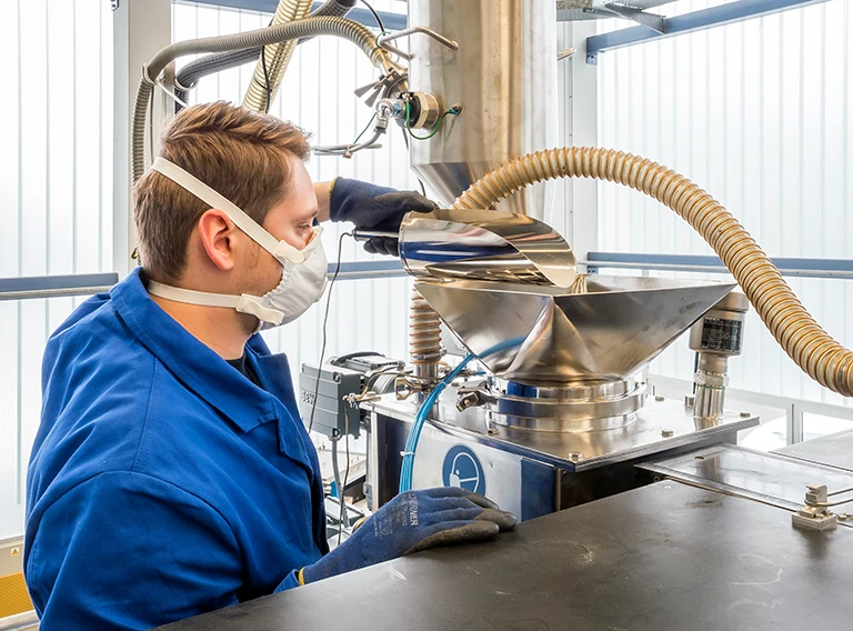 Ein Mann mit Maske, blauem Laborkittel und Handschuhen bedient industrielle Geräte in einem hellen, modernen Labor oder einer Fabrikhalle.