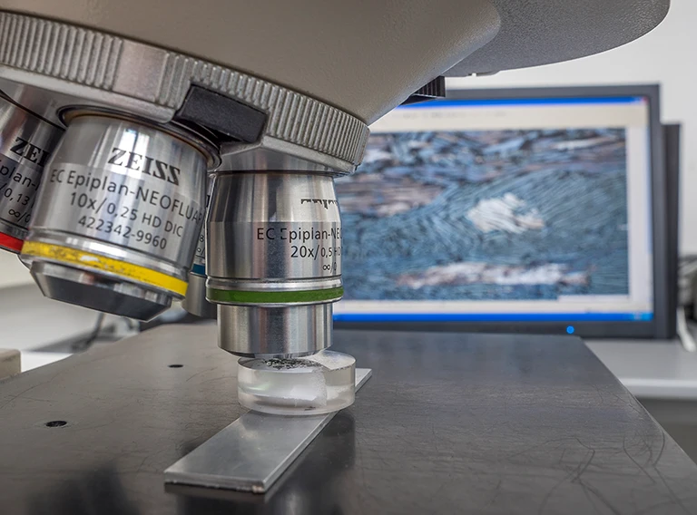 Nahaufnahme eines Mikroskopobjektivs, das einen Objektträger fokussiert, mit einem unscharfen Computerbildschirm im Hintergrund, der ein vergrößertes Bild einer blauen, strukturierten Probe anzeigt.