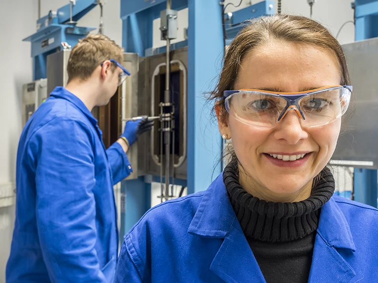 Eine Frau mit Schutzbrille und blauem Laborkittel lächelt in die Kamera, während im Hintergrund ein Mann in ähnlicher Kleidung mit Geräten in einer Laborumgebung arbeitet.