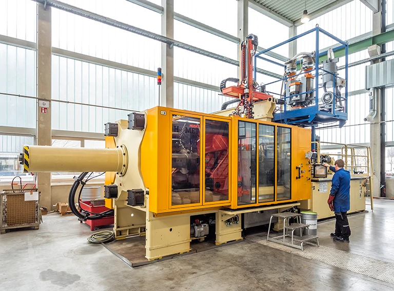 Ein Arbeiter bedient eine große gelbe Kunststoff-Spritzgießmaschine in einer geräumigen, gut beleuchteten Industrieanlage mit hohen Decken und großen Fenstern.