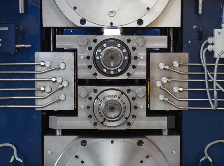 Nahaufnahme einer Industriemaschine mit Metallplatten, Bolzen, kreisförmigen Bauteilen und mehreren Metallrohren, die auf beiden Seiten miteinander verbunden sind, vor einem blauen Hintergrund.