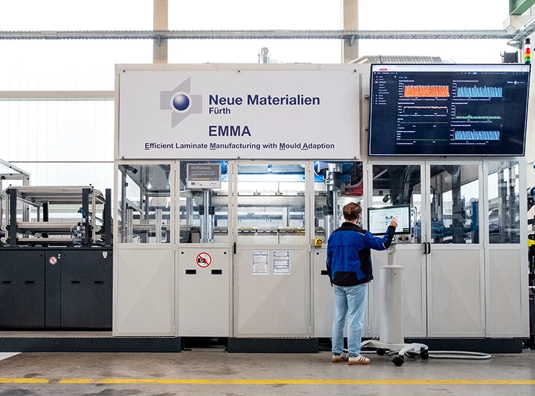 Eine Person bedient ein Bedienfeld vor einer Industriemaschine mit der Aufschrift Neue Materialien Fürth EMMA. Auf einem großen Bildschirm werden Diagramme und Daten in einer Fabrikumgebung angezeigt.