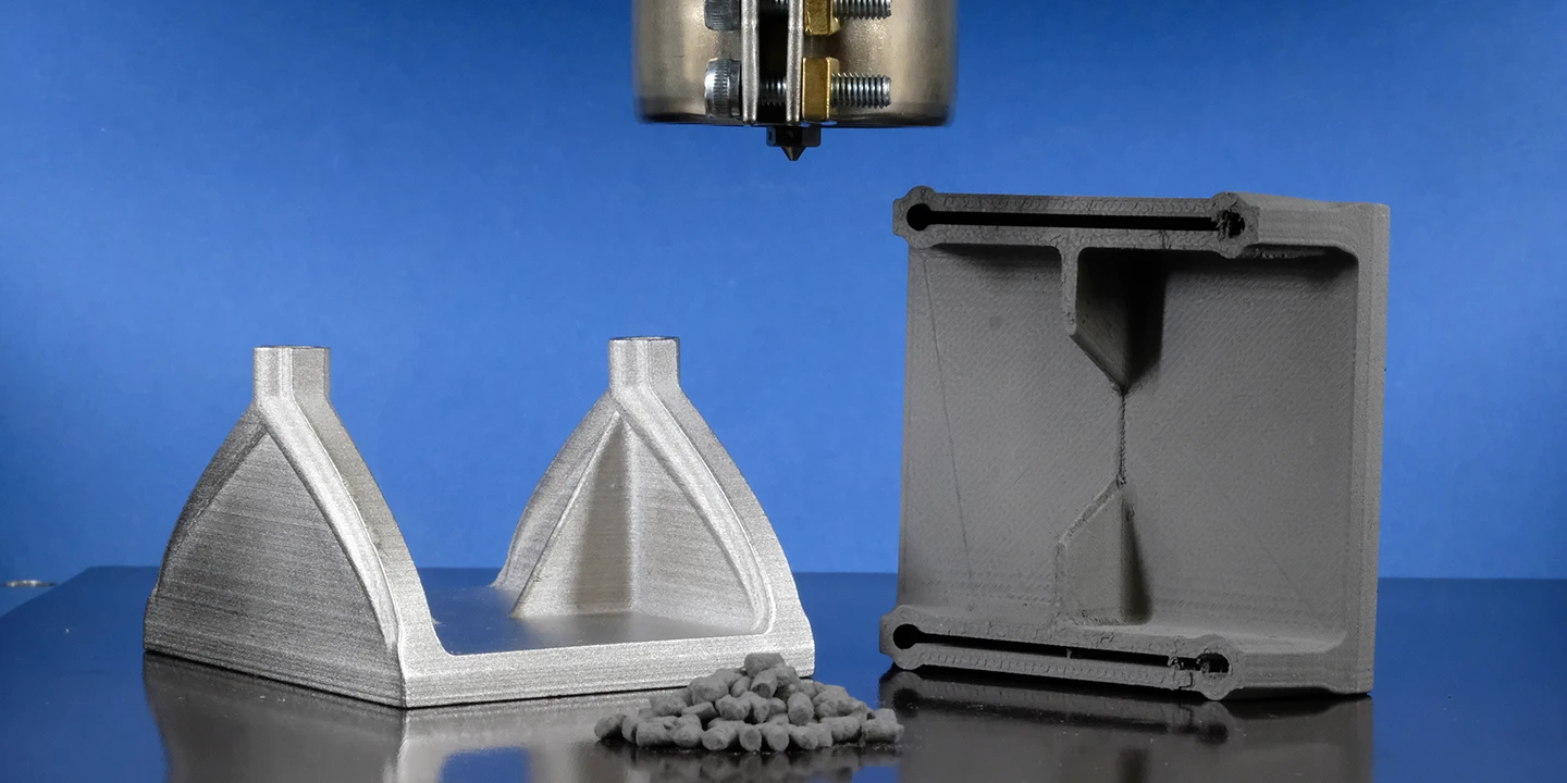 Eine 3D-Druckerdüse schwebt über einer Metallplattform mit zwei grauen 3D-gedruckten Teilen und einem kleinen Haufen Pellets vor einem blauen Hintergrund.