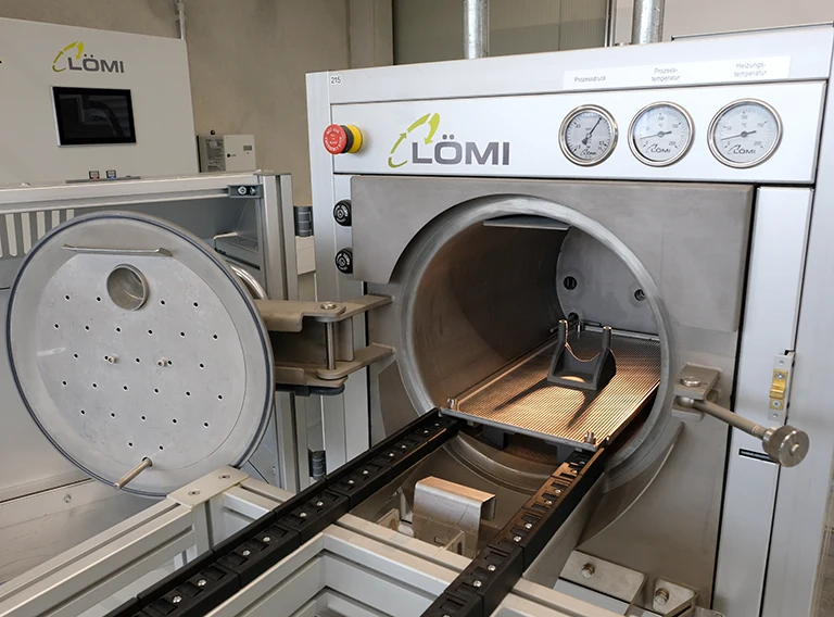 Eine Industriereinigungsmaschine mit runder, offener Tür, Innenwannen und Manometern an der Vorderseite mit der Aufschrift LÖMI wird in einer Fabrikumgebung gezeigt.