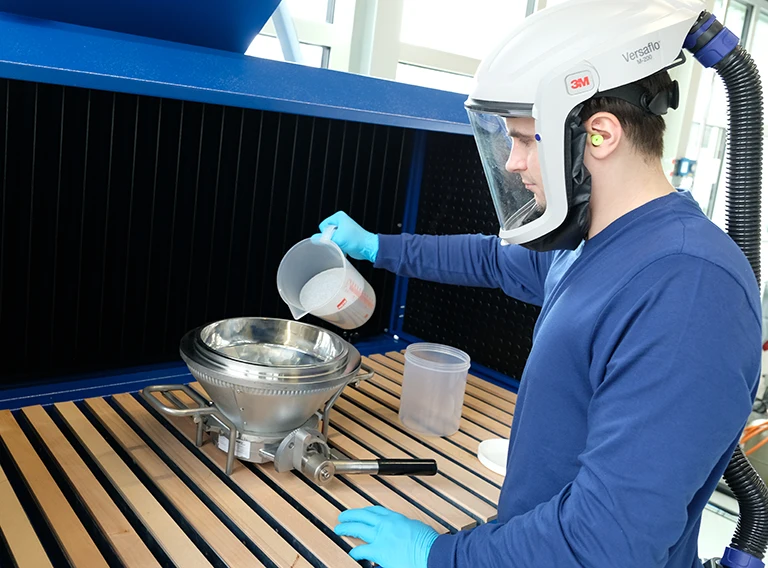 Eine Person, die Schutzhandschuhe und einen belüfteten Helm trägt, schüttet Pulver aus einem Kunststoffbehälter in einen Metalltrichter in einem Labor oder einem industriellen Arbeitsbereich.