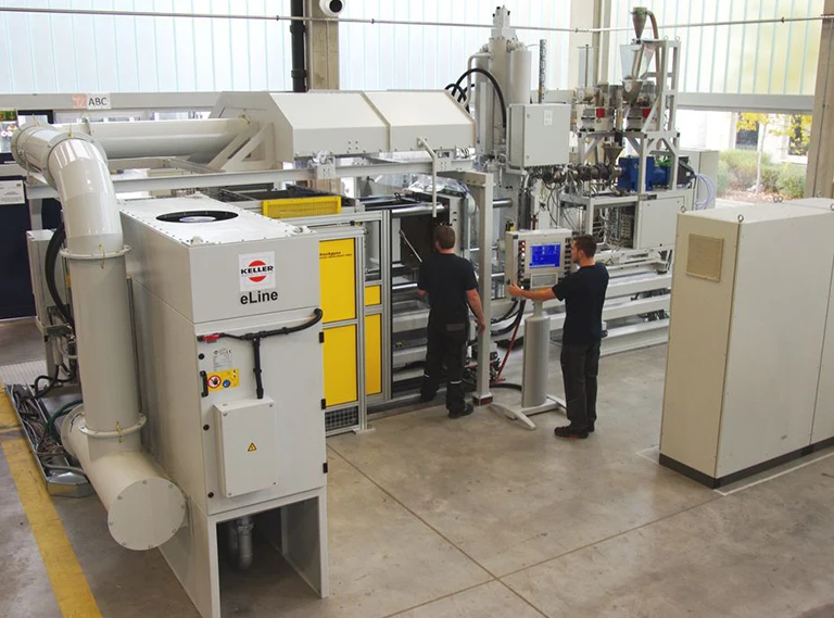 Zwei Personen stehen an einer Schalttafel und bedienen eine große Industriemaschine mit mehreren Komponenten, darunter gelbe Türen, Rohre und Entlüftungsöffnungen, in einer hellen, geräumigen Fabrikumgebung.