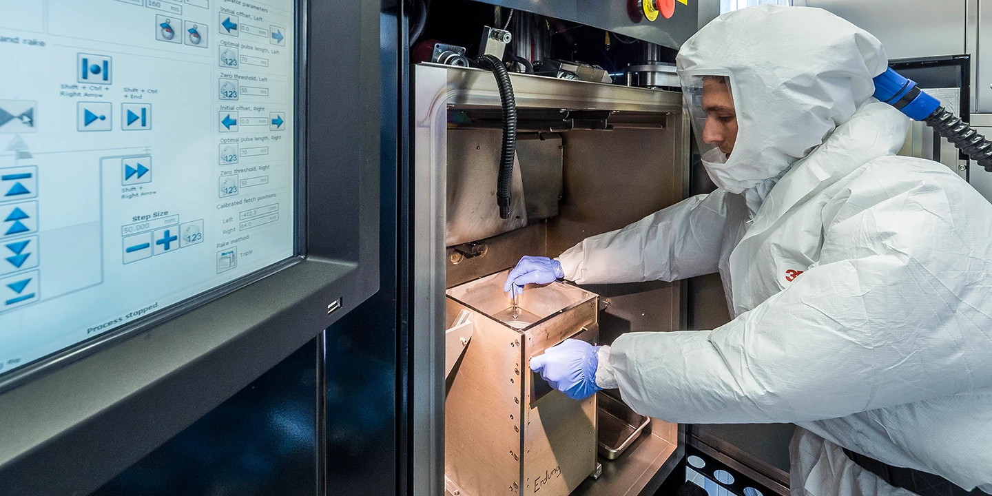Eine Person in Ganzkörperschutzanzug und Handschuhen bedient Geräte in einem Labor oder Reinraum, wobei sie mit einem Metallbehälter in der Nähe einer Maschine interagiert und die Bedienelemente auf einem Bildschirm in der Nähe sichtbar sind.