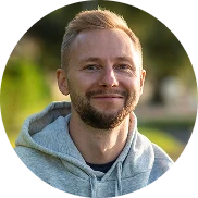 Ein lächelnder Mann mit kurzem blondem Haar und gestutztem Bart trägt einen hellgrauen Kapuzenpulli und steht im Freien mit Grün und Sonnenlicht im unscharfen Hintergrund.