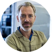 Ein Mann mit hellem Haar und Bart, der ein hellbraunes Hemd mit Kragen trägt, lächelt, während er in einem Innenraum an einem Arbeitsplatz oder in einer Industrieumgebung steht.