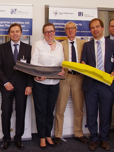 Vier professionell gekleidete Personen stehen in einem Raum und lächeln in die Kamera. Die Frau und ein Mann halten große, gebogene Industrieteile - ein graues und ein gelbes. Hinter ihnen sind technische Banner mit deutschem Text angebracht.