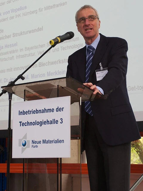 Ein Mann in Anzug und Krawatte spricht an einem Podium mit einem Mikrofon. Auf dem Podiumsschild steht Inbetriebnahme der Technologiehalle 3, Neue Materialien Fürth. Im Hintergrund ist eine Projektionsfläche mit Text zu sehen.