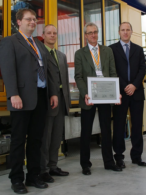 Vier Männer in Anzügen stehen in einem Haus, einer von ihnen hält eine gerahmte Urkunde in der Hand. Sie posieren für ein Foto vor einer Industrieanlage, schauen alle in die Kamera und wirken förmlich.