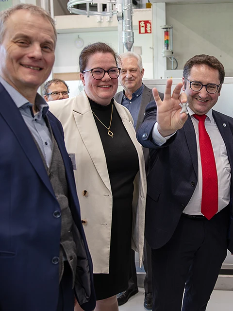 Eine Gruppe lächelnder, förmlich gekleideter Menschen steht in einem Innenraum zusammen; ein Mann in einem dunklen Anzug und mit roter Krawatte lächelt und gestikuliert mit der Hand in Richtung der Kamera.