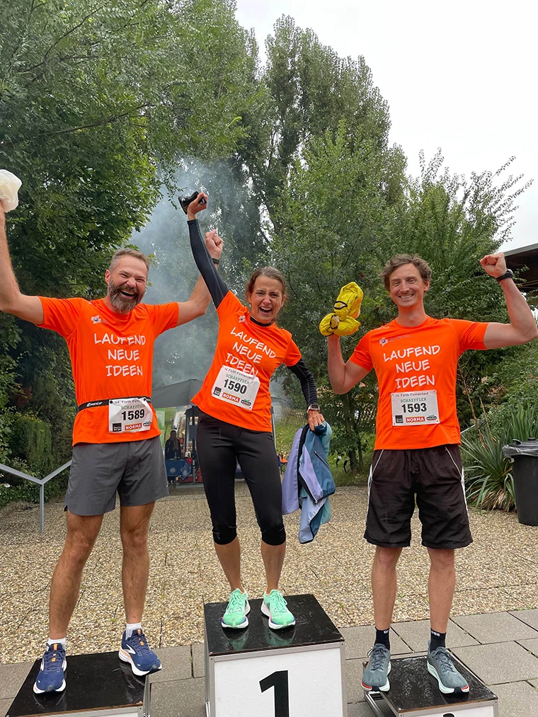 Drei Personen in passenden orangefarbenen Hemden stehen auf dem Siegerpodest, lächeln und jubeln mit zum Sieg erhobenen Armen. Sie tragen alle Startnummern und stehen im Freien mit Bäumen im Hintergrund.