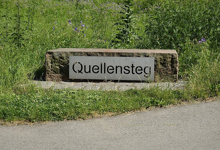 Ein steinernes Schild mit dem Wort Quellensteg steht auf einer Wiese neben einem gepflasterten Weg, mit Grünpflanzen und Wildblumen im Hintergrund.