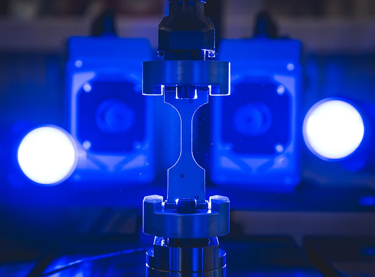 Eine Metallprobe wird in eine Zugprüfmaschine eingespannt, die von hellen blauen Lichtern beleuchtet wird. Im Hintergrund sind elektronische Geräte zu sehen, die eine Laborumgebung für Materialprüfungen darstellen.