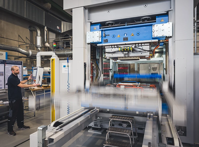 Eine Person bedient eine große Industriemaschine in einer Fabrik über ein Bedienfeld. Die Maschine ist in Bewegung, wobei unscharfe Teile die Bewegung anzeigen. Im Hintergrund sind Industrieanlagen und Rohre zu sehen.