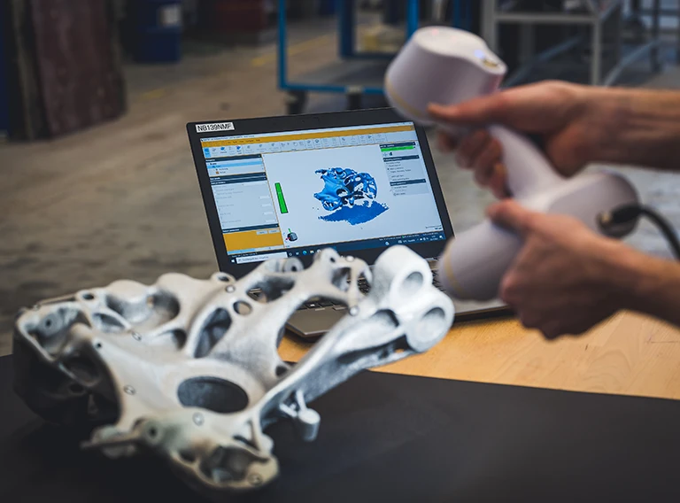 Eine Person scannt mit einem tragbaren 3D-Scanner ein komplexes, organisch geformtes Objekt, wobei die Scanergebnisse auf einem Laptop-Bildschirm im Hintergrund zu sehen sind.