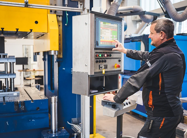 Ein Mann bedient eine große Industriemaschine über ein Bedienfeld mit Tasten und einem Touchscreen-Display in einer Fabrikumgebung. Er trägt Arbeitskleidung und scheint sich auf das Gerät zu konzentrieren.