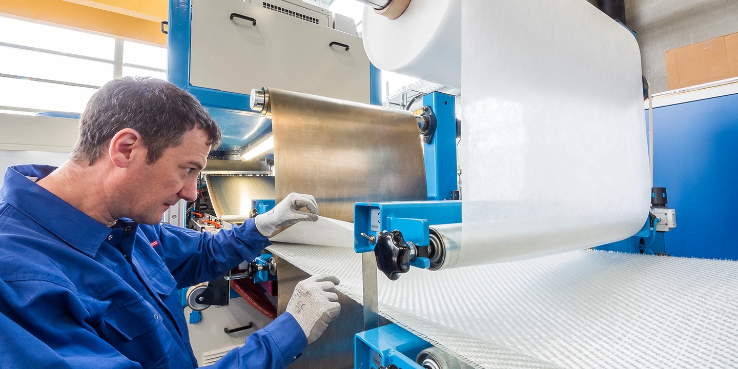 Ein Mann in blauer Arbeitskleidung und weißen Handschuhen überwacht eine große Maschine, die eine Rolle weißen Stoffs oder Materials in einer Industrieumgebung verarbeitet.