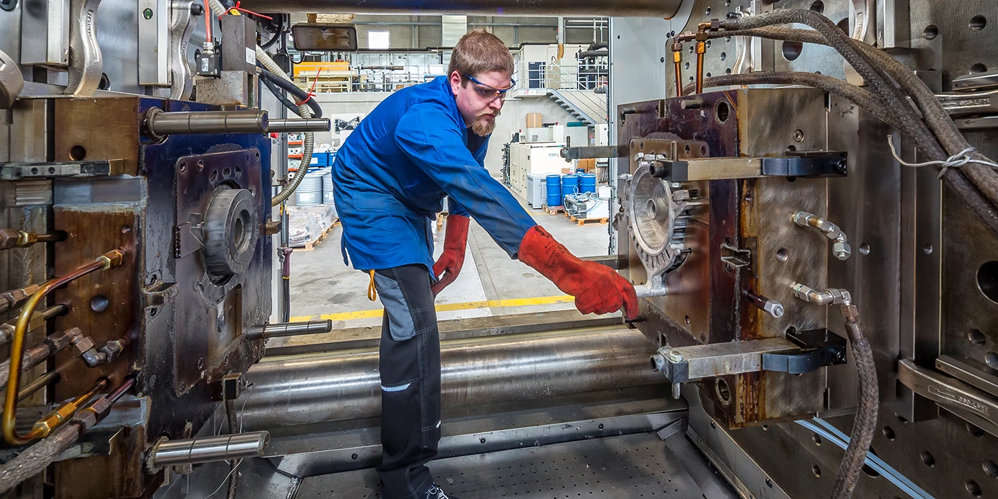 Ein Arbeiter in Schutzkleidung bedient eine große Industriemaschine in einer Fabrik und stellt mit behandschuhten Händen eine Metallform ein. Die Umgebung erscheint sauber und organisiert, mit Geräten im Hintergrund.