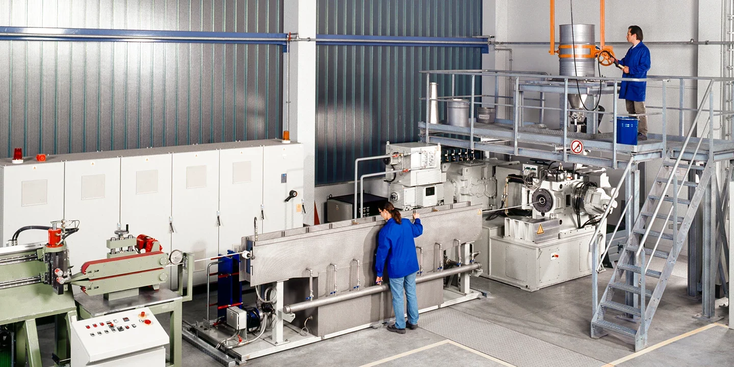 Zwei Arbeiter in blauen Kitteln bedienen Industriemaschinen in einer sauberen, modernen Fabrik. Der eine steht an einer Schalttafel, während der andere auf einer Plattform ein großes orangefarbenes Ventil dreht. Sie sind von Metallgeräten umgeben.