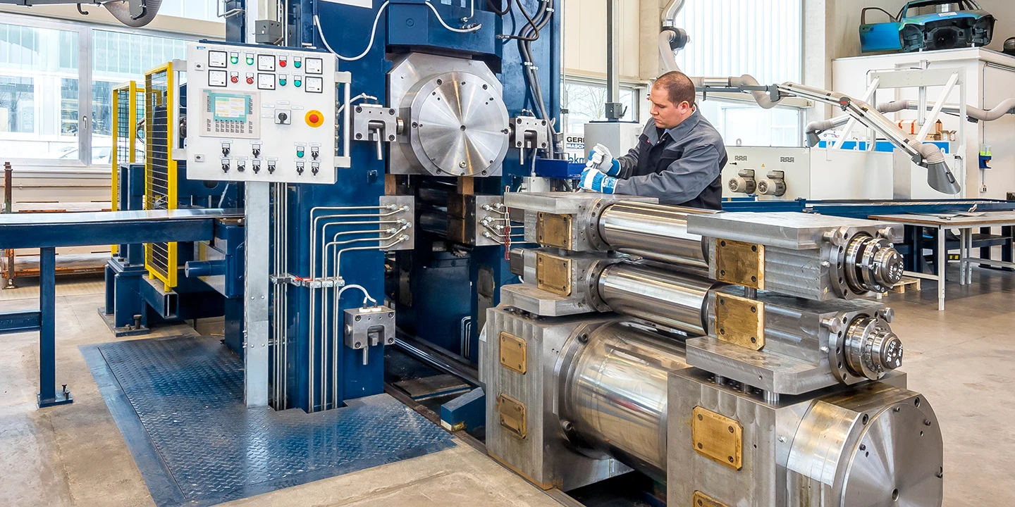 Ein Arbeiter bedient eine große Industriemaschine in einer Fabrik, in deren Nähe Metallwalzen und Zylinder gestapelt sind; in dem modernen, gut beleuchteten Arbeitsbereich sind Schalttafeln und verschiedene Maschinenteile zu sehen.