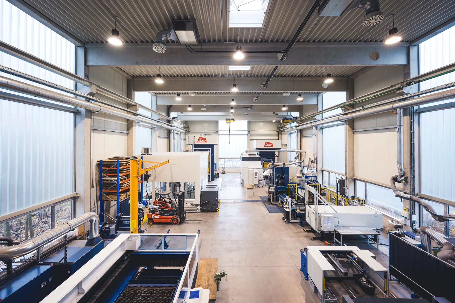 Ein geräumiges, modernes Fabrikinterieur mit verschiedenen Industriemaschinen, Metallstrukturen und Geräten, die entlang des Betonbodens angeordnet sind; große Fenster und Oberlichter erhellen den sauberen, organisierten Arbeitsbereich.