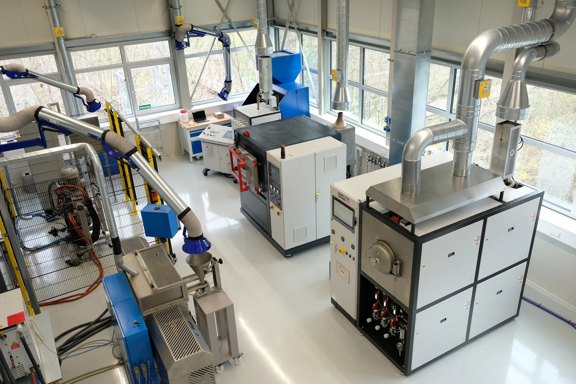 Ein modernes Industrielabor mit verschiedenen fortschrittlichen Maschinen, Lüftungsrohren und Arbeitsplätzen, alles in einem hellen Raum mit großen Fenstern und einer sauberen, organisierten Anordnung.