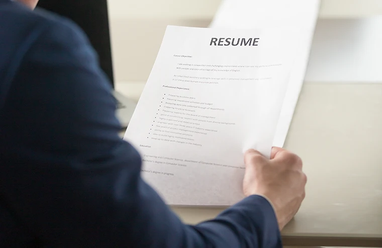 Eine Person in einem dunklen Anzug hält und liest einen gedruckten Lebenslauf an einem Schreibtisch und bereitet sich auf ein Vorstellungsgespräch oder eine Prüfung vor. Der Text des Lebenslaufs ist teilweise sichtbar, aber nicht vollständig lesbar.