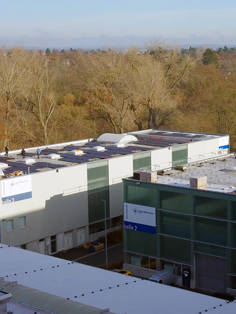 Luftaufnahme von Industriegebäuden mit Flachdächern und Schildern mit der Aufschrift halle 2 und halle 3. Mehrere Personen gehen auf den Dächern spazieren. Im Hintergrund sind blattlose Bäume und ein bewölkter Himmel zu sehen.