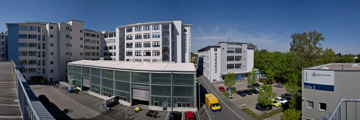 Panoramablick auf einen modernen Industriekomplex mit mehreren weißen mehrstöckigen Gebäuden, Glasfassaden, geparkten Fahrzeugen, Bäumen und einem klaren blauen Himmel.