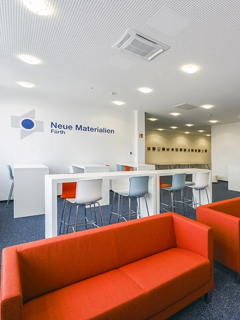 Eine moderne Büro-Lounge mit orangefarbenen Sofas, weißen Stehtischen und grauen und weißen Barhockern. An der Wand hängen das Logo und der Text Neue Materialien Fürth. Der Raum ist gut beleuchtet und hat einen blauen Teppich.