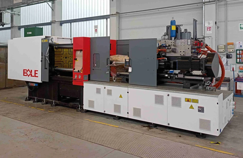 Eine industrielle Spritzgießmaschine mit roten, weißen und grauen Platten steht in einer Fabrik, umgeben von Sicherheitsschildern und -ausrüstung.