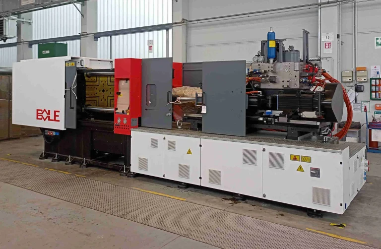 Eine industrielle Spritzgießmaschine mit roten, weißen und grauen Platten steht in einer Fabrik, umgeben von Sicherheitsschildern und -ausrüstung.