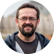 Ein Mann mit kurzen braunen Haaren, einer Brille und einem Bart lächelt. Er trägt ein graues T-Shirt und einen blauen Kapuzenpulli und steht im Freien vor einem unscharfen Holzzaun im Hintergrund.