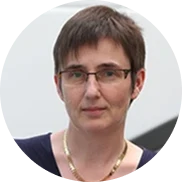 Eine Person mit kurzen braunen Haaren, die eine Brille, ein dunkles Oberteil und eine goldene Halskette trägt, schaut mit neutralem Blick in die Kamera. Der Hintergrund ist unscharf und in hellen Farben gehalten.