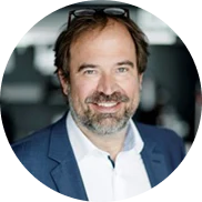 Ein lächelnder Mann mit braunem Haar und Bart trägt einen blauen Blazer und ein weißes Hemd. Er hat eine Brille auf dem Kopf und ist in einem Innenraum mit unscharfem Hintergrund abgebildet.