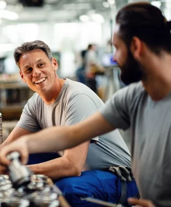Zwei Männer in legerer Arbeitskleidung lächeln und unterhalten sich in einer hell erleuchteten Industrie- oder Werkstattumgebung, in der im Hintergrund Werkzeuge und Ausrüstung zu sehen sind.