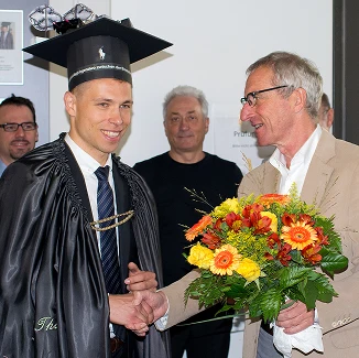 Ein junger Mann mit Abschlussmütze und Talar schüttelt einem älteren Mann mit einem Strauß orangefarbener und gelber Blumen die Hand, während im Hintergrund drei Personen lächelnd stehen.