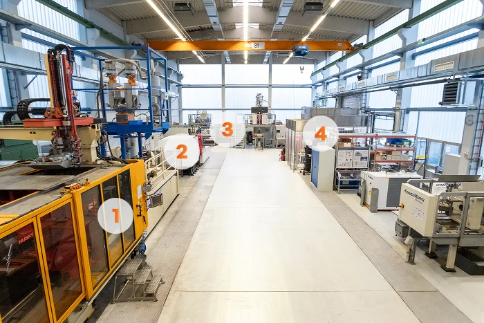 Eine geräumige, gut beleuchtete Industriehalle mit nummerierten Arbeitsplätzen und verschiedenen großen Maschinen und Geräten auf beiden Seiten eines breiten Mittelgangs.