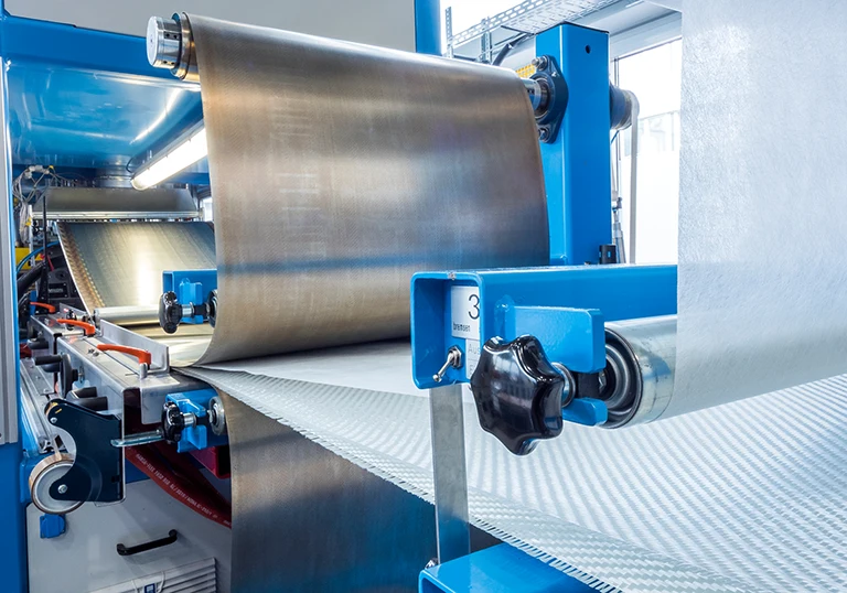 Nahaufnahme einer Industriemaschine, die Stoff- oder Materialrollen verarbeitet, mit blauen Metallteilen und großen Walzen, die das Material durch die Anlage in einer Produktionsumgebung führen.