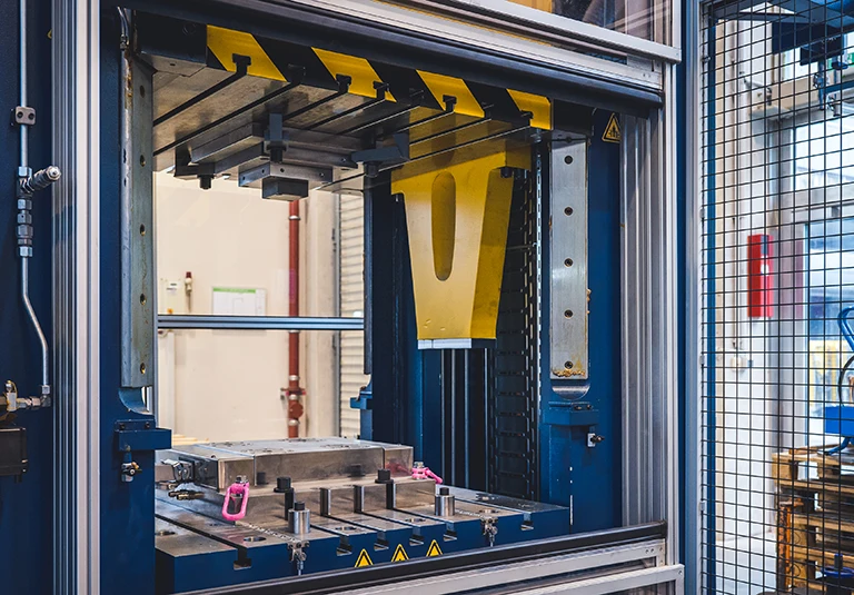 Eine Nahaufnahme einer großen Industriepresse in einer Fabrik, die Metallteile, Sicherheitsschranken und Bedienfelder zeigt, mit Werkzeugen und Teilen, die auf der Grundplatte angeordnet sind.